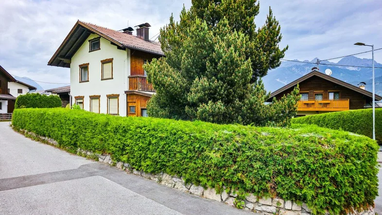 Traditionelles Haus mit weißer Fassade, Holzverkleidung und gepflegter Hecke in alpiner Umgebung.