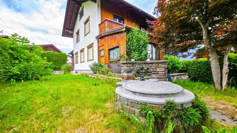Gartenbereich mit Rasenfläche, einem dekorativen Brunnen und dem Haus im Hintergrund, umgeben von Bäumen.
