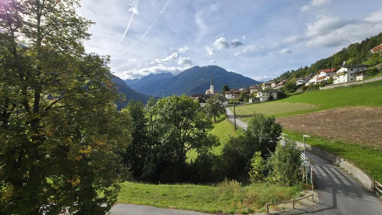 Malerischer Ausblick auf ein grünes Tal mit Bergen, einem Dorf und weiten Feldern unter blauem Himmel.