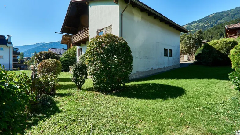 Seitenansicht eines traditionellen Hauses mit gepflegtem Garten und Bergblick unter blauem Himmel.