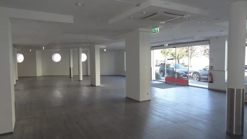 Großer, leerer Gewerberaum mit grauem Laminatboden, weißen Wänden und großen Schaufenstern zur Straße.