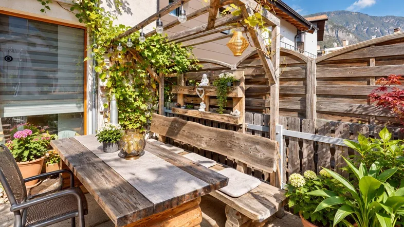 Rustikale Terrasse mit Holztisch, Bänken und üppiger Begrünung unter einer Pergola.