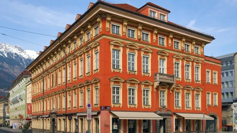 Historisches Gebäude mit auffälliger roter Fassade und gelben Verzierungen, umgeben von einer städtischen Straße mit Bergblick.