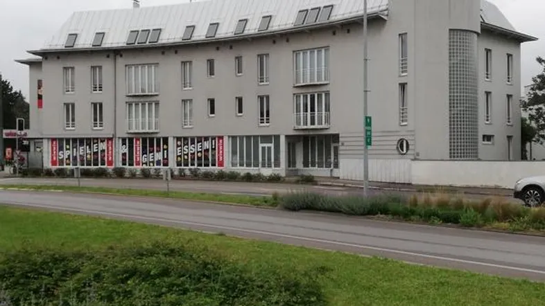 Graues Mehrfamilienhaus mit moderner Architektur und Geschäften im Erdgeschoss, umgeben von Grünflächen.