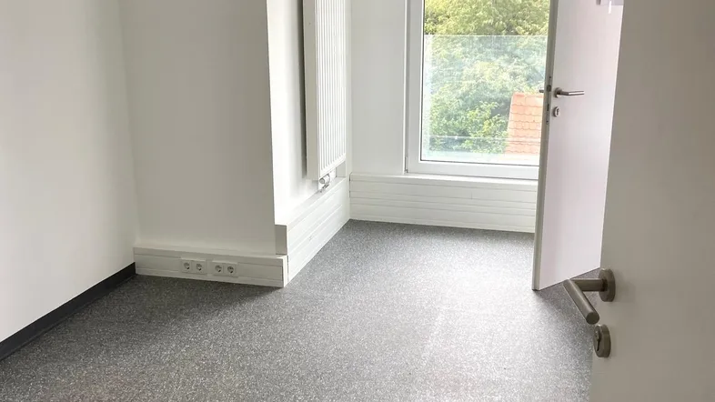 Heller, leerer Raum mit grauem Vinylboden, weißen Wänden und einem Fenster mit Blick ins Grüne.