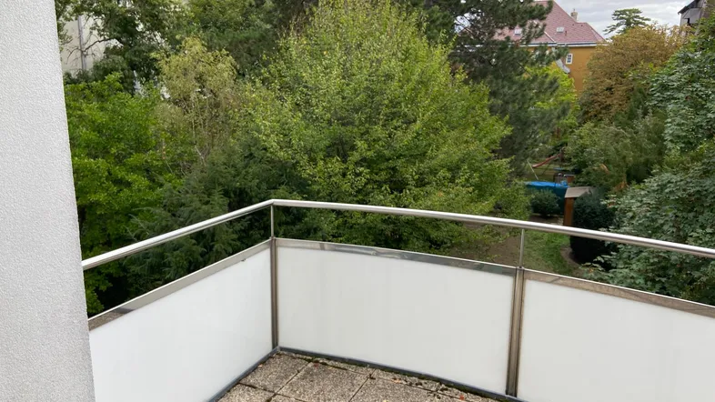 Geräumiger Balkon mit Fliesenboden und Blick ins Grüne, umgeben von Bäumen und anderen Gebäuden.