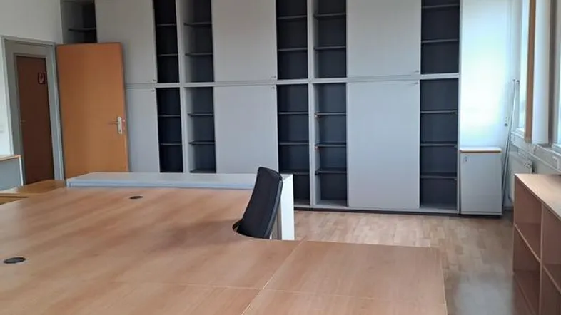 Großes Büro mit hellem Holzschreibtisch, Bürostuhl und integrierten Regalschränken. Der Raum verfügt über Holzfußboden und Deckenbeleuchtung.