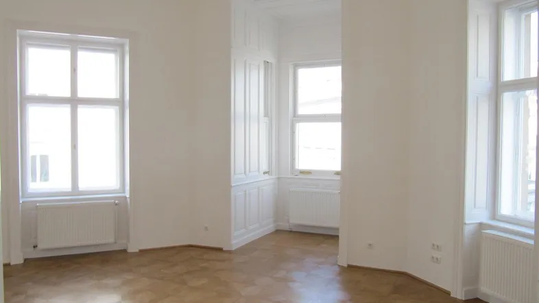 Helles Zimmer mit Fischgrätparkett und hohen Fenstern, das viel Tageslicht hereinlässt.