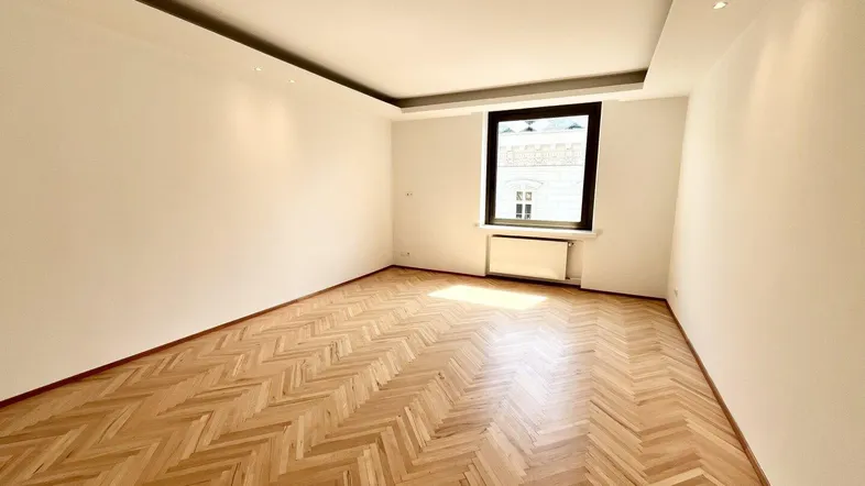 Helles, leeres Zimmer mit Fischgrätparkettboden und einem Fenster, das Tageslicht hereinlässt.