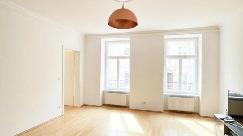 Helles Wohnzimmer mit Parkettboden, hohen Decken und Stuckelementen, durchflutet von natürlichem Licht.