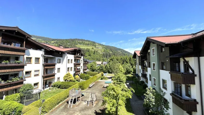 Blick auf die Wohnanlage mit Spielplatz und grünen Hügeln im Hintergrund unter blauem Himmel.