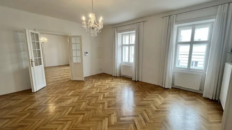 Geräumiges Wohnzimmer mit Fischgrätparkett, hohen Fenstern und einem eleganten Kronleuchter.
