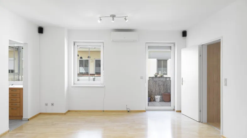 Helles, leeres Wohnzimmer mit Parkettboden, Klimaanlage und Zugang zum Balkon.