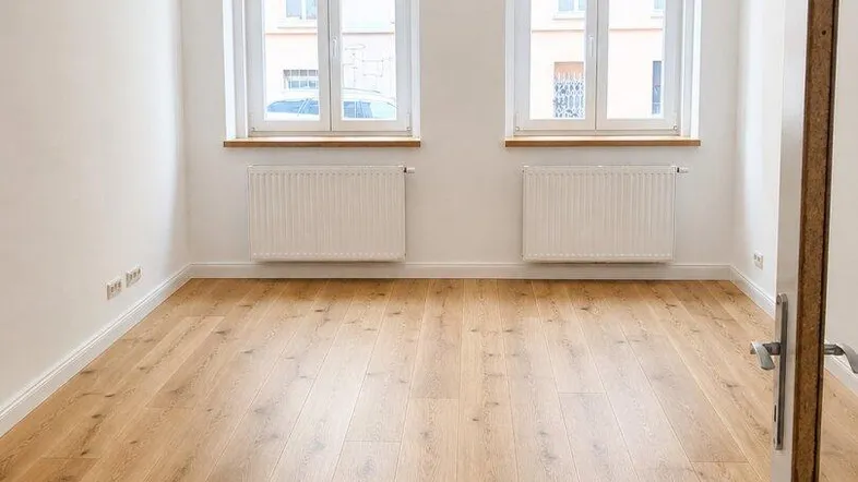 Heller, leerer Raum mit zwei Fenstern, Holzfußboden und weiß gestrichenen Wänden.