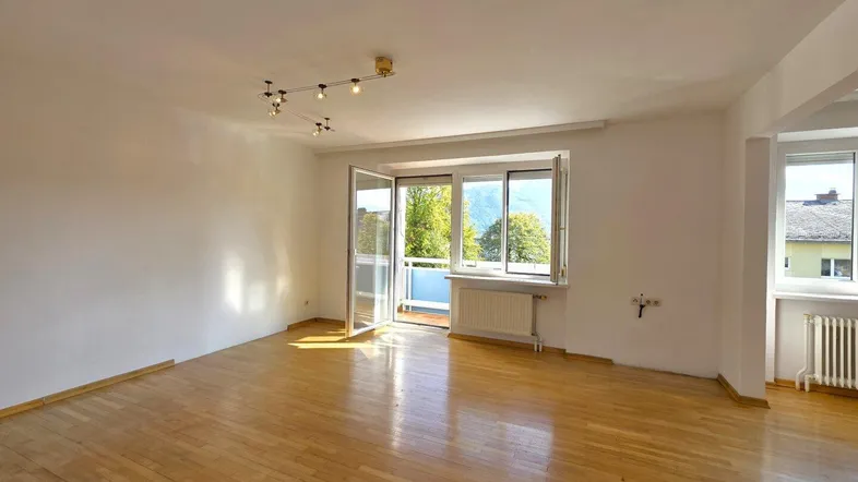 Helles, leeres Wohnzimmer mit Parkettboden, weißen Wänden und Zugang zu einem Balkon.