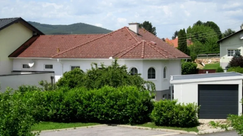 Weißes Einfamilienhaus mit rotem Ziegeldach, umgeben von grünen Büschen und einem Garagengebäude.