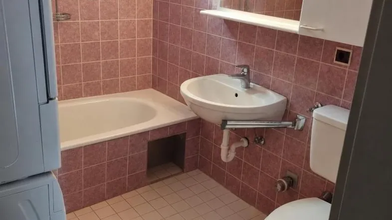 Badezimmer mit rosa Fliesen, Badewanne, Waschbecken und Toilette, ausgestattet mit Waschmaschine.