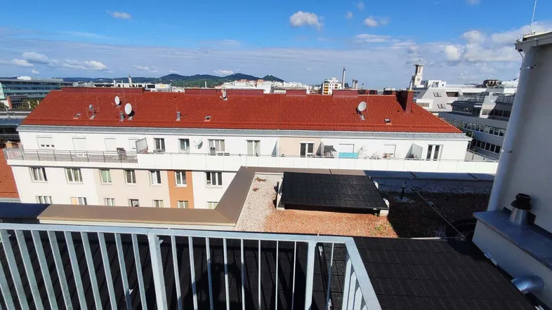 Blick über die Dächer der Stadt mit blauem Himmel und fernen Hügeln von einem Balkon.