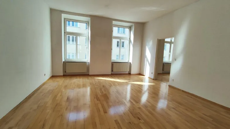 Helles Zimmer mit glänzendem Parkettboden und zwei großen Fenstern, die viel Tageslicht hereinlassen.