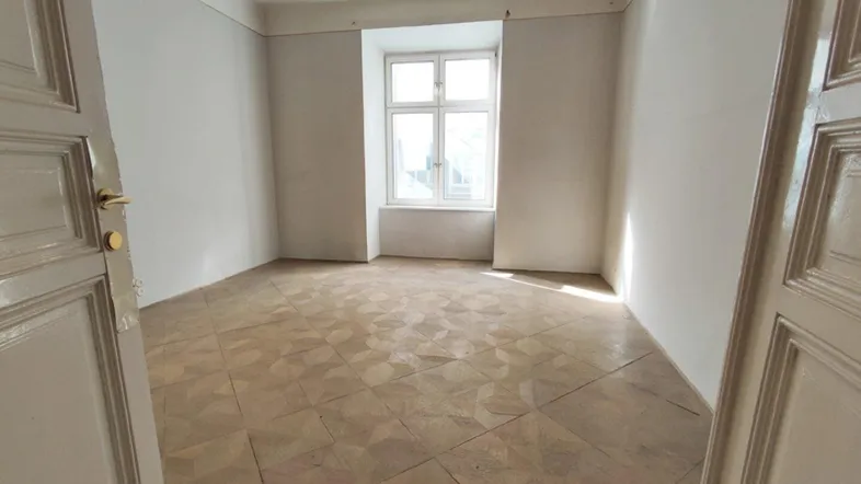 Helles Zimmer mit Fischgrätparkettboden und einem großen Fenster, bereit zur Renovierung.