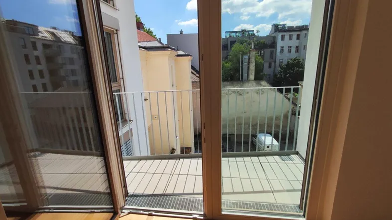 Blick aus einem hellen Raum mit Holzboden auf einen kleinen Balkon und die umliegenden Gebäude.