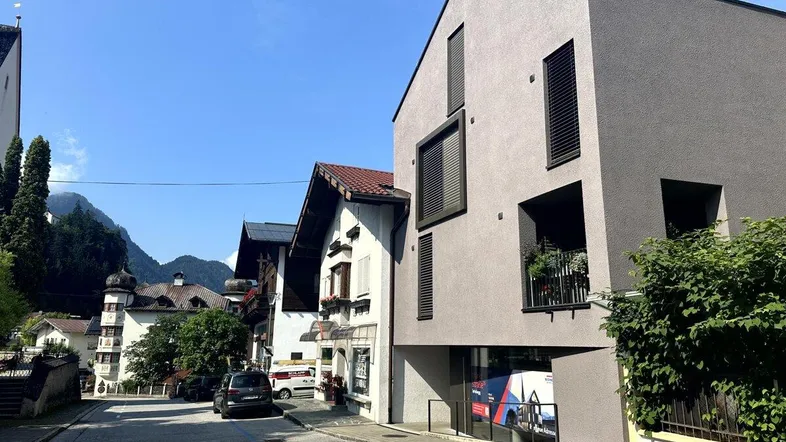 Modernes Gebäude mit grauer Fassade und dunklen Fensterläden in einer belebten Straße mit Bergblick.