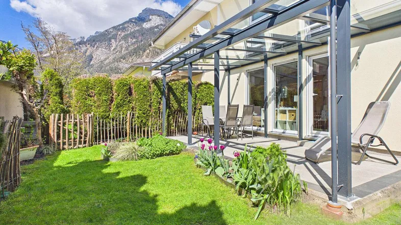 Überdachte Terrasse mit Gartenmöbeln, Rasenfläche und Bergblick im Hintergrund.