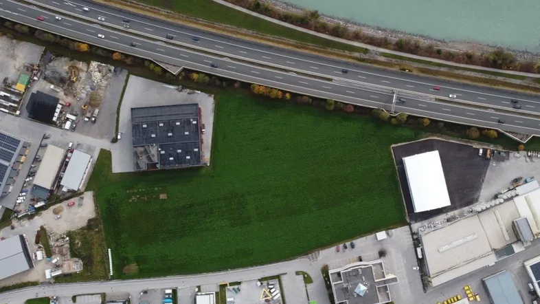 Luftaufnahme eines großen grünen Grundstücks neben einer Autobahn und einem Fluss, umgeben von Gewerbegebäuden.