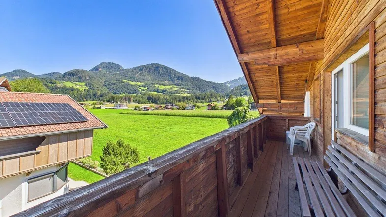 Geräumiger Holzbalkon mit Sitzbank und weitem Blick auf die umliegende Berglandschaft und grüne Wiesen.