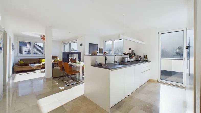 Helle, offene Wohnküche mit moderner Kücheninsel, Essbereich und Zugang zum Balkon, alles auf elegantem Fliesenboden.