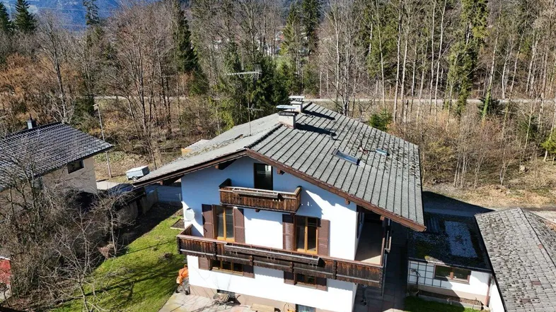 Luftaufnahme des Hauses mit Satteldach und Balkonen, umgeben von Bäumen und Bergen.