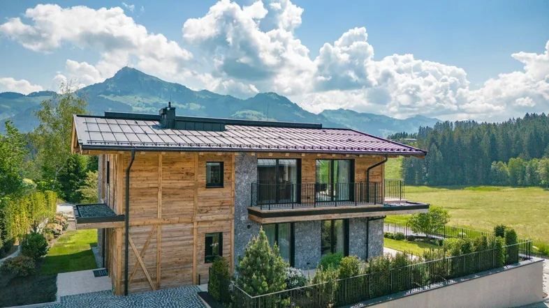 Modernes Holzhaus mit Steinfassade und Balkonen vor alpiner Bergkulisse und grüner Landschaft.