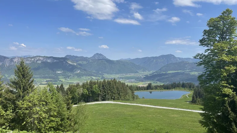 Weitläufige Berglandschaft mit grünem Tal, Wäldern und einem kleinen See unter blauem Himmel.