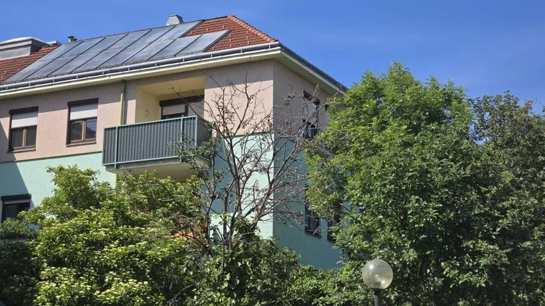 Mehrfamilienhaus mit Balkon, Photovoltaikanlage auf dem Dach und umgebender Vegetation.