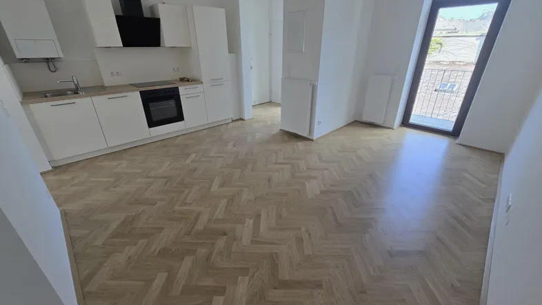 Heller offener Wohnbereich mit moderner Einbauküche, Fischgrätparkett und Zugang zu einem Balkon.