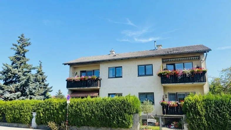 Zweistöckiges Gebäude mit blühenden Balkonen und gepflegter Hecke unter blauem Himmel.