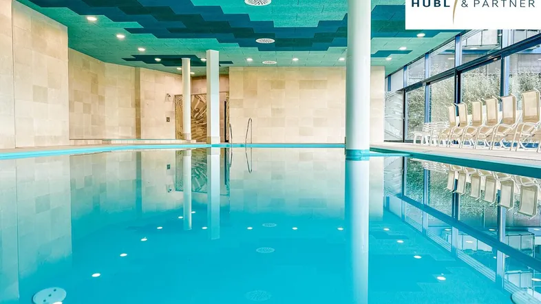 Modernes Hallenbad mit großem Schwimmbecken, Liegestühlen und Blick ins Grüne.
