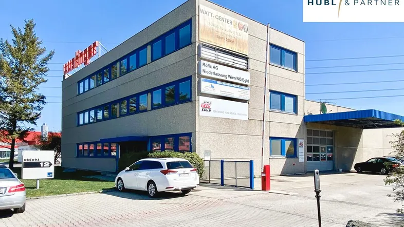 Mehrstöckiges Gewerbegebäude mit blauen Fensterrahmen und Parkplätzen davor unter blauem Himmel.