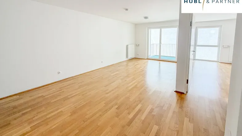 Helles, leeres Wohnzimmer mit Parkettboden und Zugang zu einem Balkon.