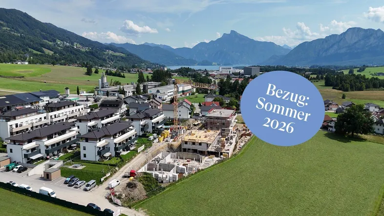Luftaufnahme einer Baustelle mit neuen Wohngebäuden, See- und Bergblick im Hintergrund.