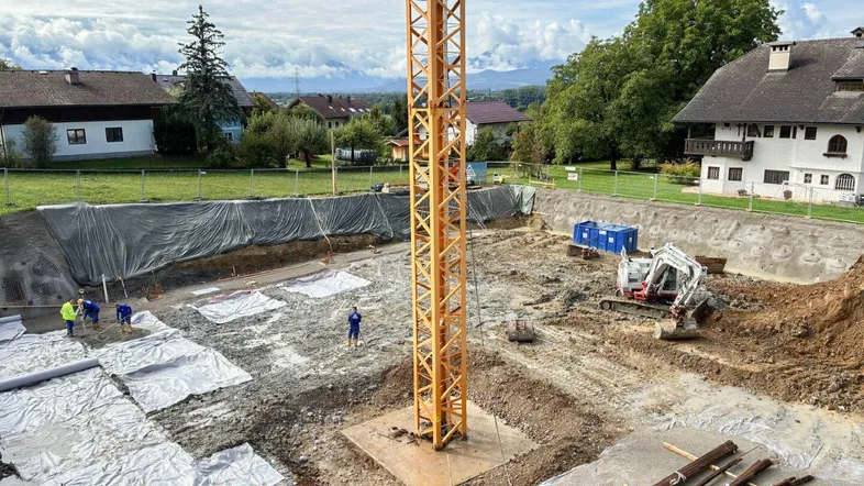 Baufortschritt einer Baustelle mit ausgehobener Grube, Kran und Arbeitern, im Hintergrund Berge.