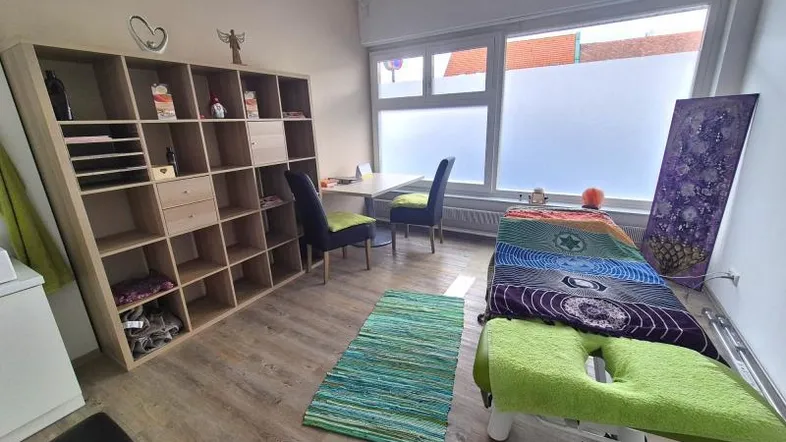 Heller Behandlungsraum mit großem Fenster, Holzregal und Liegefläche, ideal für Therapien oder Beratungen.