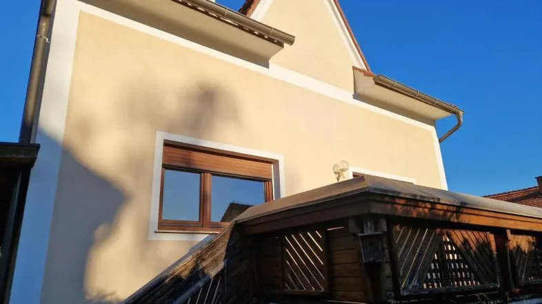 Ansicht der Hausfassade mit einem erhöhten Holzbalkon und einem Fenster mit Rollladen.