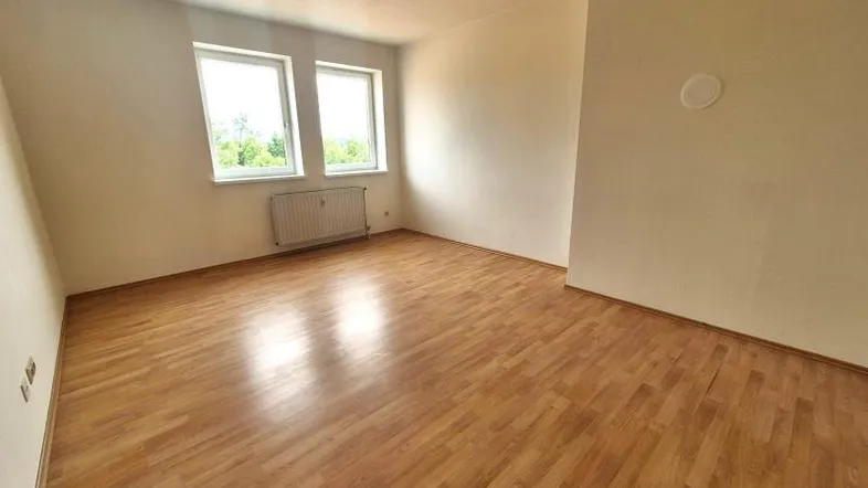 Helles Zimmer mit Laminatboden, beigen Wänden und zwei Fenstern mit Blick ins Grüne.