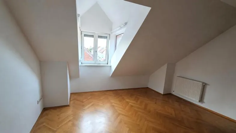 Helles Dachzimmer mit schrägen Wänden, Fenster und Fischgrätparkettboden, ideal als Schlaf- oder Arbeitszimmer.
