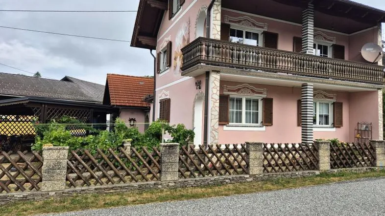 Traditionelles Haus mit rosafarbener Fassade, Holzbalkon und Fensterläden, umgeben von einem Lattenzaun.