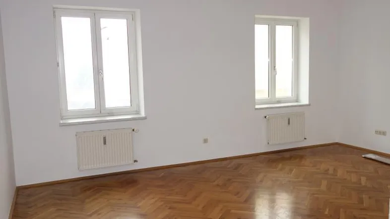 Helles Zimmer mit weiß gestrichenen Wänden, zwei Fenstern und klassischem Fischgrätparkettboden.