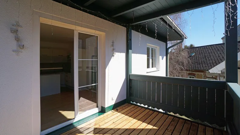 Heller Balkon mit Holzboden und dunklem Geländer, zugänglich durch eine Glasschiebetür.
