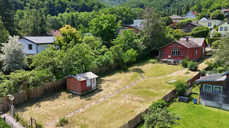 Luftaufnahme eines Grundstücks mit rotem Haus, Schuppen und weitläufiger Grünfläche in hügeliger Landschaft.