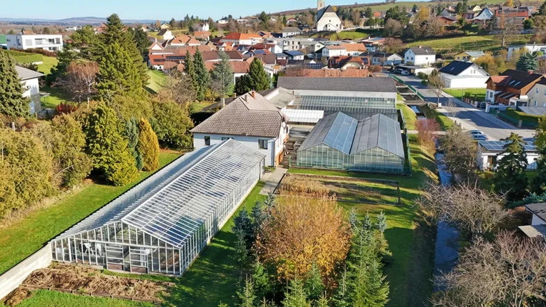 Weitläufige Luftaufnahme des Anwesens mit Gewächshäusern, Wohnhaus und Blick auf das umliegende Dorf mit Kirche.